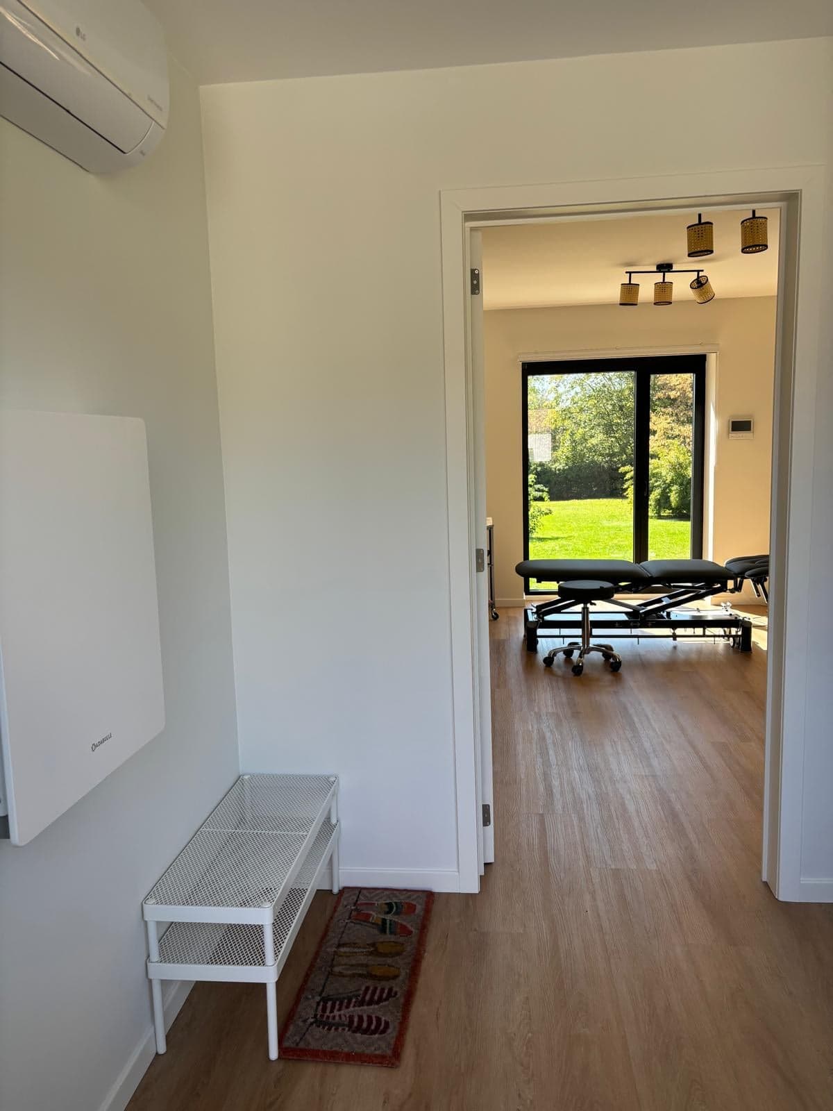Entrée du cabinet avec un banc blanc, la porte ouverte donne sur la salle de soin et la baie vitrée