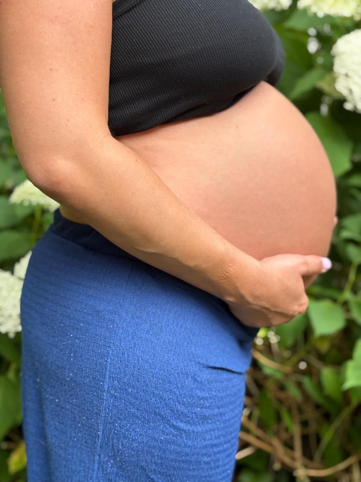 Ventre de femme enceinte de profil, une main posée dessous, devant des hortensias